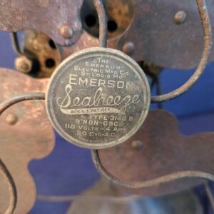 Peut inclure: Gros plan d'un ventilateur &eacute;lectrique vintage Emerson Seabreeze. Le ventilateur comporte un embl&egrave;me circulaire avec le nom de la marque et les sp&eacute;cifications. Les pales et la cage sont en m&eacute;tal avec un aspect vieilli. Le ventilateur est un mod&egrave;le non oscillant.