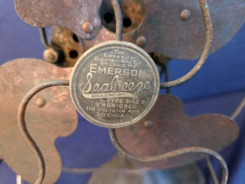 Peut inclure: Gros plan d'un ventilateur &eacute;lectrique vintage Emerson Seabreeze. Le ventilateur comporte un embl&egrave;me circulaire avec le nom de la marque et les sp&eacute;cifications. Les pales et la cage sont en m&eacute;tal avec un aspect vieilli. Le ventilateur est un mod&egrave;le non oscillant.