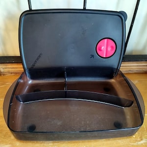 May include: A black plastic divided lunch container with a red button on the lid. The lid has the text "Tupperware" printed on it.