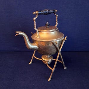 May include: Antique brass tea kettle with a black handle and a stand. The kettle has a curved spout and a lid with a small knob. The stand has three legs and a circular base. The kettle is approximately 10 inches tall.