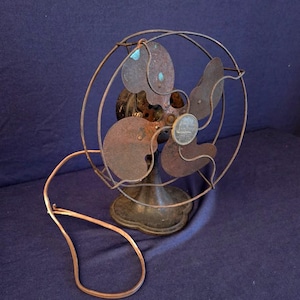 Peut inclure: Un ancien ventilateur &eacute;lectrique &agrave; l'aspect rouill&eacute; et vieilli. Le ventilateur a quatre pales et une cage de protection en fil de fer. Le socle est orn&eacute; et le cordon d'alimentation est de couleur cuivre. Les composants m&eacute;talliques du ventilateur montrent des signes d'usure et d'&acirc;ge.