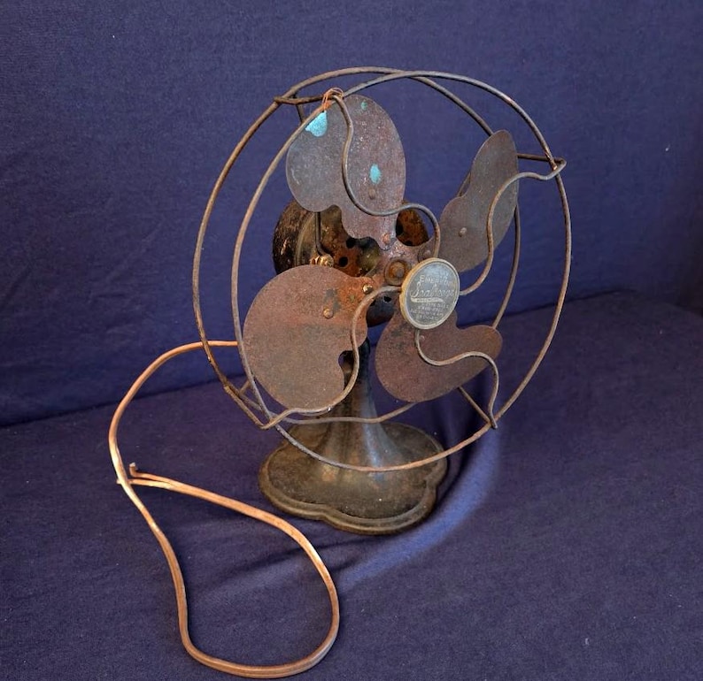 Peut inclure: Un ancien ventilateur &eacute;lectrique &agrave; l'aspect rouill&eacute; et vieilli. Le ventilateur a quatre pales et une cage de protection en fil de fer. Le socle est orn&eacute; et le cordon d'alimentation est de couleur cuivre. Les composants m&eacute;talliques du ventilateur montrent des signes d'usure et d'&acirc;ge.