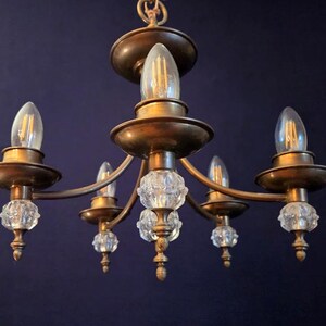 May include: A vintage chandelier with five lights. The fixture has a brass finish and features clear, faceted crystal accents. The bulbs are teardrop-shaped. The chandelier is suspended against a dark blue background.