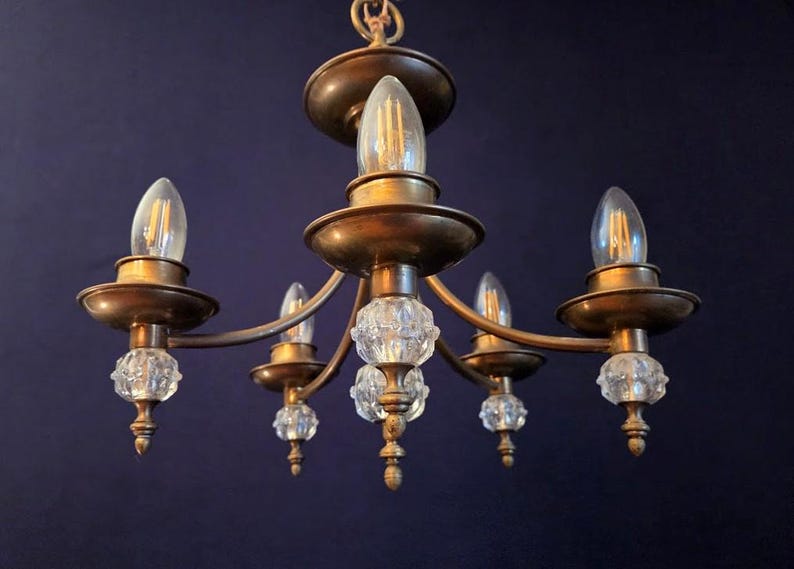 May include: A vintage chandelier with five lights. The fixture has a brass finish and features clear, faceted crystal accents. The bulbs are teardrop-shaped. The chandelier is suspended against a dark blue background.