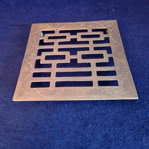 May include: A square, metallic decorative object with a geometric openwork design. The object has a series of rectangular and horizontal cutouts, with a decorative scalloped edge. The object is set against a dark blue background.