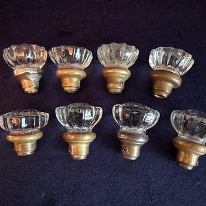 May include: Eight clear glass cabinet knobs with brass-colored bases are arranged on a dark blue surface. The knobs have a faceted, flower-like design. The brass bases show signs of age.