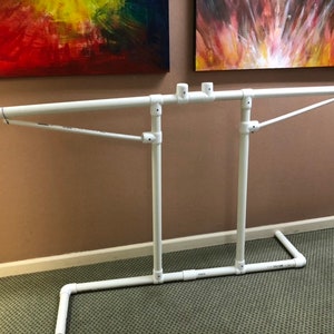 Może przedstawiać: Biała rama z rur PVC z dwoma poziomymi prętami i ukośnym wspornikiem. Rama jest wolnostojąca i wydaje się być projektem DIY.