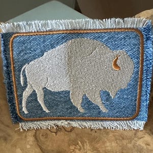 May include: A rectangular denim patch featuring a light gray embroidered bison silhouette in the center. The patch is outlined with a copper-colored stitched border and finished with frayed, white fabric edges on the top and bottom sides.