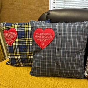 May include: Two plaid throw pillows made from repurposed shirts. Each pillow features a red heart-shaped appliqué with embroidered text. One pillow is blue and yellow plaid, the other is gray and blue plaid.