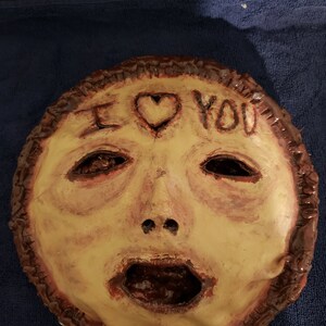 May include: A handmade ceramic mask with a distressed, painted finish. The mask features a human face with a large, open mouth and a heart with the words "I love you" written above it.
