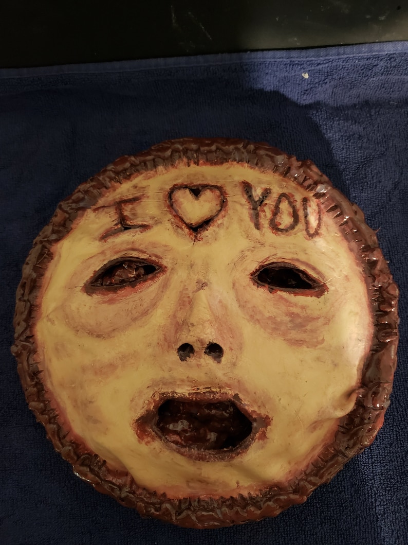 May include: A handmade ceramic mask with a distressed, painted finish. The mask features a human face with a large, open mouth and a heart with the words "I love you" written above it.