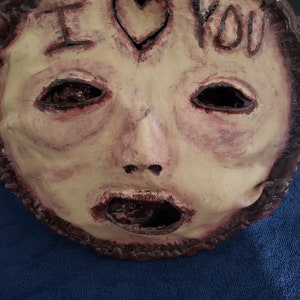 May include: A creepy, realistic-looking mask with a human face. The mask is made of a flesh-toned material and has a large, open mouth. The eyes are black and hollow, and the nose is slightly crooked. The mask has a heart drawn on the forehead with the words "I love you" written above it.
