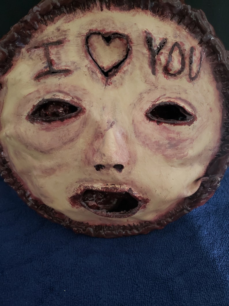 May include: A creepy, realistic-looking mask with a human face. The mask is made of a flesh-toned material and has a large, open mouth. The eyes are black and hollow, and the nose is slightly crooked. The mask has a heart drawn on the forehead with the words "I love you" written above it.