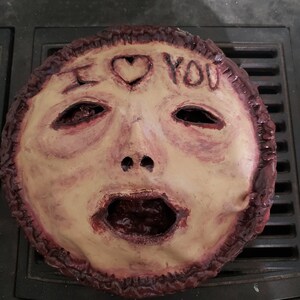 May include: A creepy, realistic-looking mask made of paper mache. The mask is painted in shades of brown and red, and features a human face with a large, open mouth. The words "I LOVE YOU" are written on the forehead of the mask, with a heart symbol in between the words.