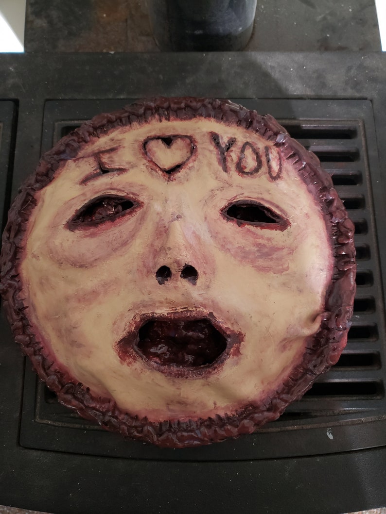 May include: A creepy, realistic-looking mask made of paper mache. The mask is painted in shades of brown and red, and features a human face with a large, open mouth. The words "I LOVE YOU" are written on the forehead of the mask, with a heart symbol in between the words.