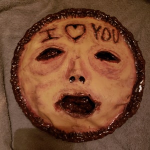 May include: A creepy, hand-painted mask with a human face, open mouth, and the words "I love you" written on the forehead. The mask is made of a brown material and has a textured surface.