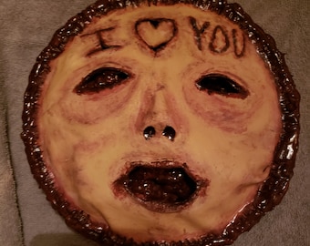 Könnte beinhalten: Eine gruselige, handbemalte Maske mit einem menschlichen Gesicht, offenem Mund und den Worten "Ich liebe dich" auf der Stirn. Die Maske ist aus braunem Material und hat eine strukturierte Oberfläche.