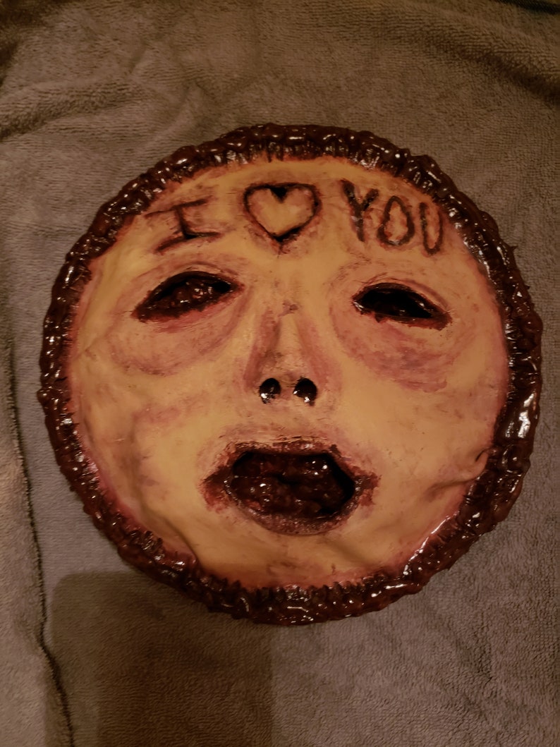 May include: A creepy, hand-painted mask with a human face, open mouth, and the words "I love you" written on the forehead. The mask is made of a brown material and has a textured surface.