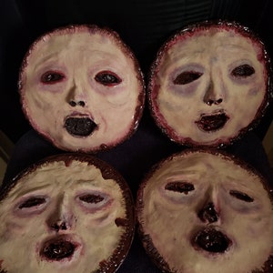 May include: Four round ceramic masks with painted faces. The masks are painted in shades of white, pink, and red. The masks have a distressed look and appear to be made of clay.