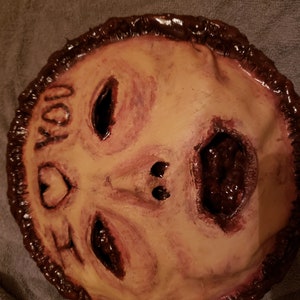 May include: A handmade mask with a distressed, flesh-toned face and a dark brown border. The mask has a large, open mouth with a dark red interior and two black eyes. The text "I LOVE YOU" is written on the mask in black.