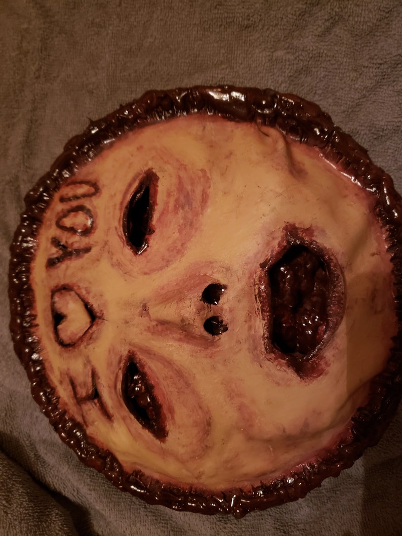 May include: A handmade mask with a distressed, flesh-toned face and a dark brown border. The mask has a large, open mouth with a dark red interior and two black eyes. The text "I LOVE YOU" is written on the mask in black.