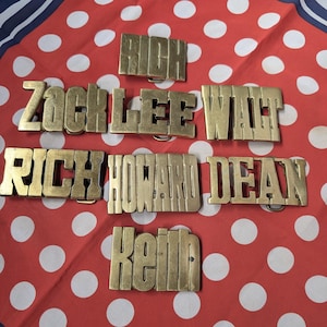 May include: A collection of brass belt buckles, each spelling out a different name: Rich, Zack, Lee, Walt, Howard, Dean, and Keith. The buckles are displayed on a red and white polka dot background.