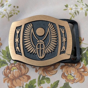 May include: A gold-toned belt buckle with a black and gold design of an Egyptian winged scarab beetle. The buckle is attached to a black leather belt strap.
