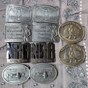 May include: A collection of vintage metal belt buckles. The buckles feature various designs, including eagles, the NRA logo, and text such as "Defender of Firearms Freedom" and "NRA Life Member". The buckles are in shades of silver and bronze.