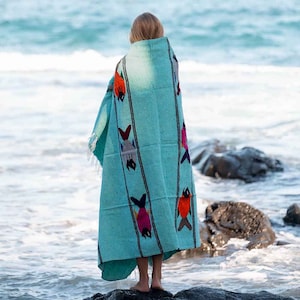 May include: A person standing on a rocky beach, wrapped in a turquoise blue blanket with colourful fish designs. The blanket has a fringe border.