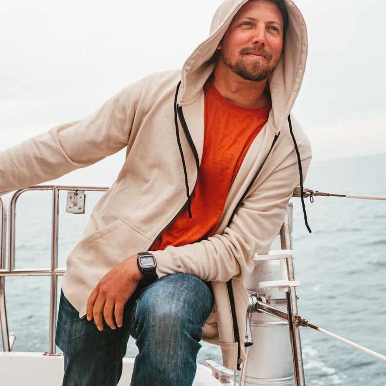 K&ouml;nnte beinhalten: Beige Kapuzen-Sweatshirt mit schwarzem Rei&szlig;verschluss und einem leuchtend orangefarbenen T-Shirt darunter. Die Person tr&auml;gt eine blaue Jeans und eine schwarze Uhr. Der Hintergrund zeigt ein Gew&auml;sser und einen bew&ouml;lkten Himmel.