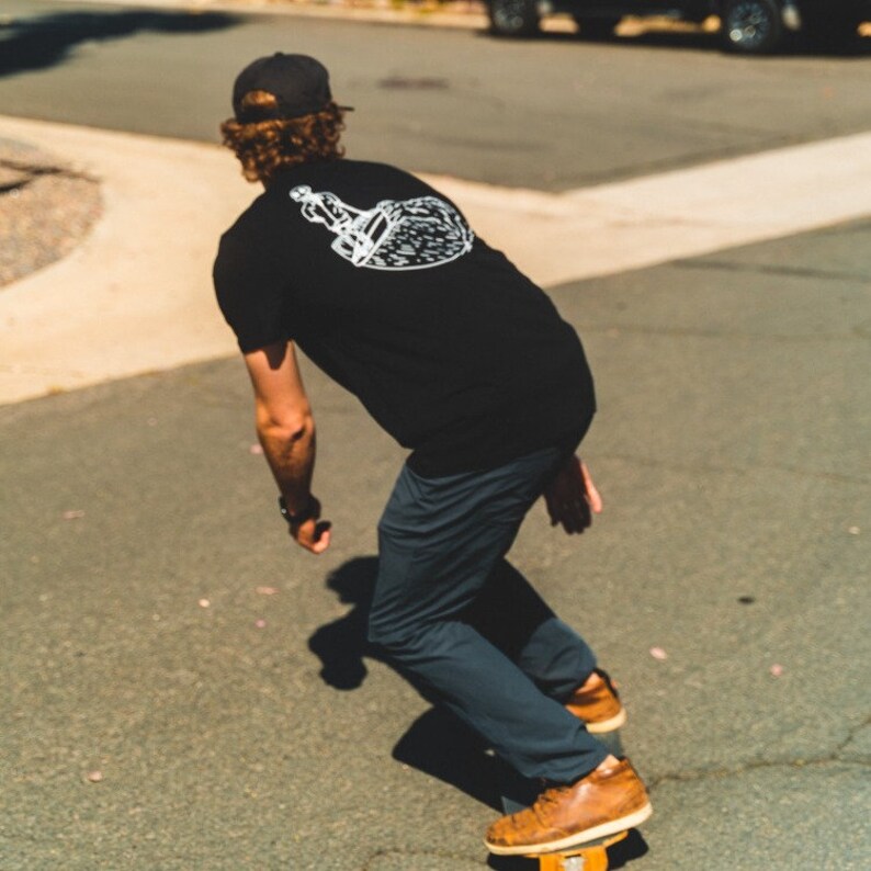 May include: A person skateboarding on a paved road, wearing a black t-shirt with a white graphic on the back, dark pants, a black cap, and brown shoes. The skateboard is yellow and orange.