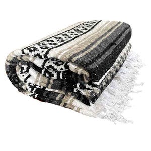 Puede incluir: Una manta mexicana de rayas negras, blancas y beige con flecos. La manta está doblada y tiene un patrón tradicional.