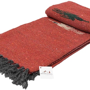 Pu&ograve; includere: Una coperta rossa piegata con frange grigio scuro. La coperta ha una trama tessuta e un disegno in verde e bianco. Un'etichetta con il testo "OPEN ROAD GOODS" &egrave; allegata.