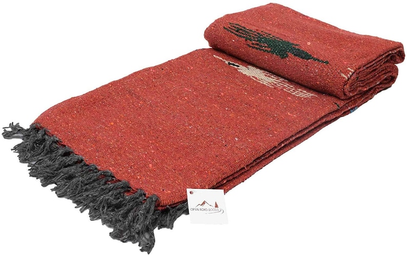 Pu&ograve; includere: Una coperta rossa piegata con frange grigio scuro. La coperta ha una trama tessuta e un disegno in verde e bianco. Un'etichetta con il testo "OPEN ROAD GOODS" &egrave; allegata.