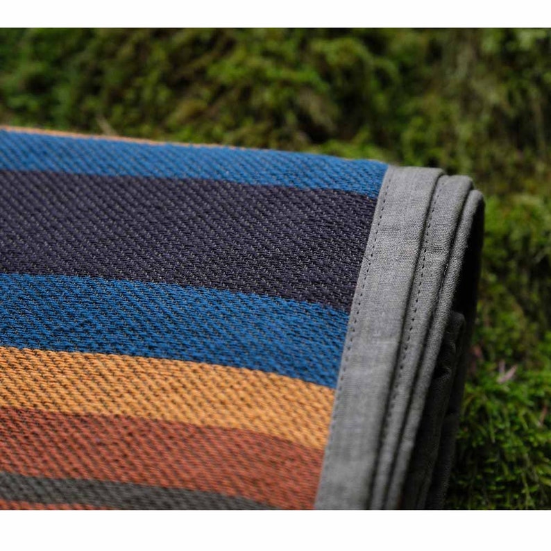 May include: Folded outdoor blanket with horizontal stripes in shades of blue, orange, and brown. The blanket has a gray border and is made of a woven fabric. The blanket is resting on a bed of green moss.