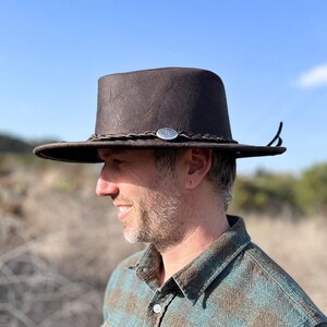 May include: A brown leather cowboy hat with a braided band and a silver concho. The hat has a wide brim and a tall crown.