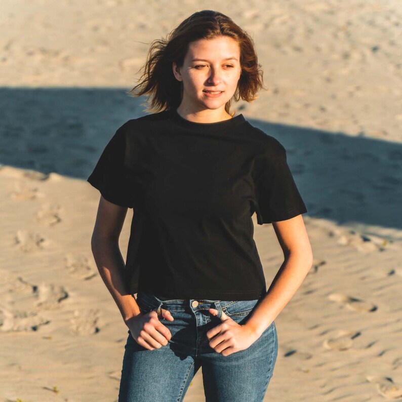 K&ouml;nnte beinhalten: Ein schwarzes T-Shirt mit Rundhalsausschnitt und eine blaue Jeans. Die Person steht an einem Sandstrand mit den H&auml;nden in den Taschen. Das T-Shirt hat kurze &Auml;rmel und eine klassische Passform. Die Jeans ist mittelblau.