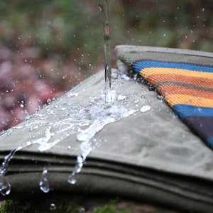 May include: A stack of olive green canvas blankets with a colorful striped blanket on top. Water is pouring onto the blankets, creating splashes and droplets. The striped blanket features orange, blue, and brown stripes.