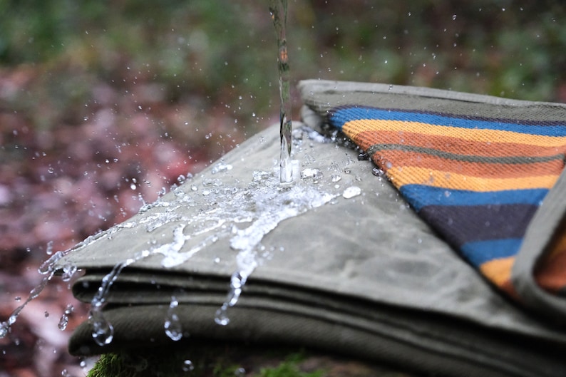 May include: A stack of olive green canvas blankets with a colorful striped blanket on top. Water is pouring onto the blankets, creating splashes and droplets. The striped blanket features orange, blue, and brown stripes.