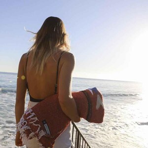 May include: A woman standing on a wooden deck overlooking the ocean, holding a red and white striped woven blanket. The blanket has a fringe border and a white tag with the text "Made in Mexico".