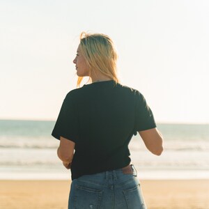 K&ouml;nnte beinhalten: Eine Person tr&auml;gt ein schwarzes T-Shirt und blaue Jeans an einem Strand. Die Person steht mit dem R&uuml;cken zum Betrachter und blickt aufs Meer. Der Himmel ist hell, und das Meer ist im Hintergrund zu sehen. Die Jeans sind im Used-Look.