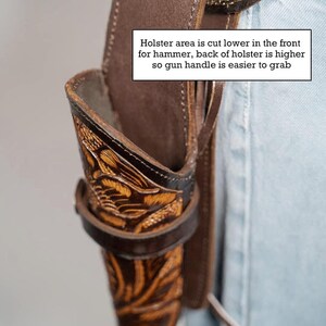 Pode incluir: Um coldre de couro marrom com detalhes florais intrincados. O coldre &eacute; projetado para ser usado em um cinto e tem um corte mais baixo na frente para facilitar o acesso ao martelo de uma arma de fogo. "Holster area is cut lower in the front for hammer, back of holster is higher so gun handle is easier to grab" est&aacute; escrito na imagem.