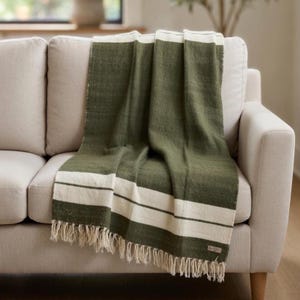 May include: A green and white striped throw blanket draped over a beige sofa. The blanket features a dark green base with wide white stripes and fringe along the bottom edge. The sofa has a neutral color and a simple design.
