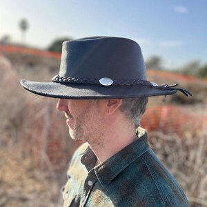 May include: A black fedora-style hat with a wide brim. The hat features a braided band with silver-colored embellishments and a leather tie detail. The hat is worn outdoors, suggesting a casual or outdoor theme.