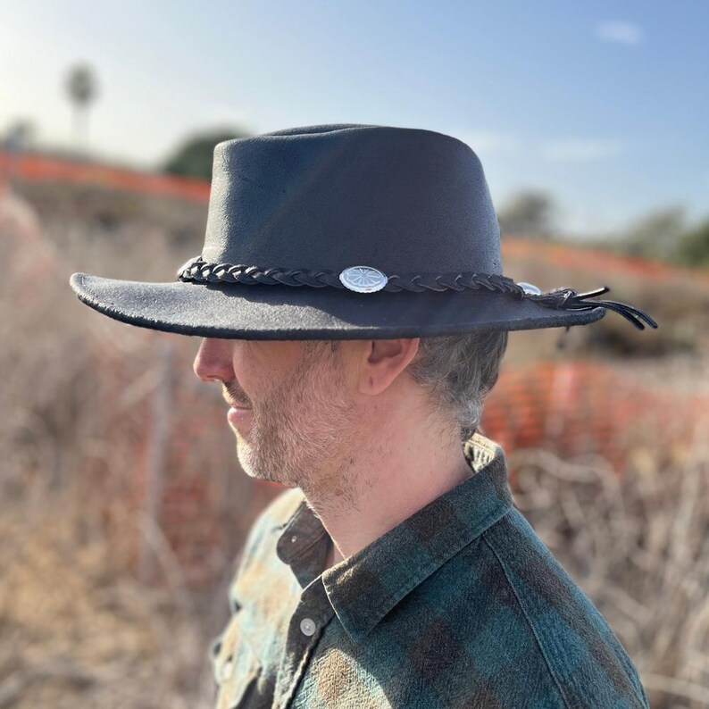 May include: A black fedora-style hat with a wide brim. The hat features a braided band with silver-colored embellishments and a leather tie detail. The hat is worn outdoors, suggesting a casual or outdoor theme.