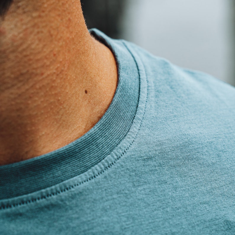 May include: A close-up shot of a teal crew neck t-shirt. The neckline is ribbed with detailed stitching. The fabric has a soft, textured appearance. The image focuses on the collar and shoulder area of the shirt.
