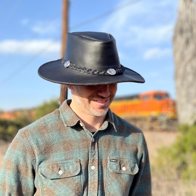 May include: Black leather hat with a wide brim, braided band, and silver-toned embellishments. The hat has a classic cowboy hat shape. The hat is worn by a person wearing a plaid shirt.
