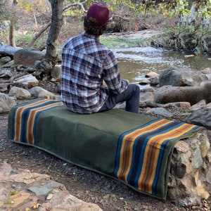 May include: A green outdoor bench with a colorful striped blanket draped over it. The blanket features stripes of blue, yellow, and brown. The bench is made of stone and is located near a river.