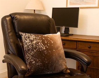 May include: A brown and white cowhide-patterned decorative pillow rests on a dark brown leather office chair. The pillow has a soft, textured appearance. The chair is in a home office setting, with a desk, computer, and lamp visible in the background.