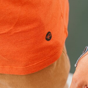 K&ouml;nnte beinhalten: Nahaufnahme eines orangefarbenen T-Shirts mit einem schwarzen Logo auf der unteren linken Seite. Das Logo ist ein stilisiertes "WO"-Design. Das Shirt besteht aus einem weichen, strukturierten Stoff.
